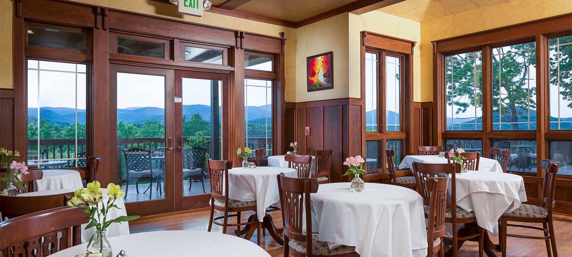 An interior dining room with dark stained woodword on a soft yellow wall pallate. Draped tables with seating for two each, fill the scene.