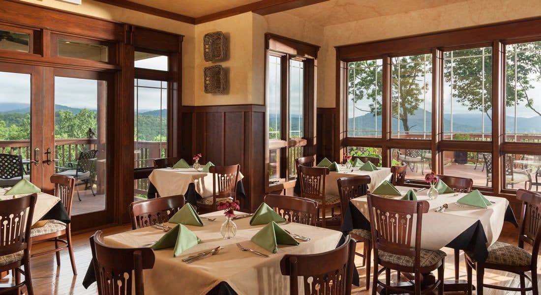 Dining room with white table cloths and green napkins with windows on two sides with view of the mountains.