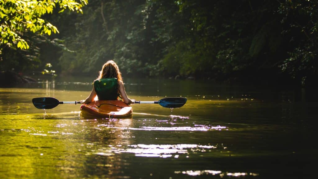 A woman holding paddles in a kayak on a partially shaded body of water.