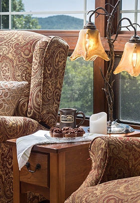 Wingback chairs flank a small table with antique lamp with scenic mountain view outside the windows in the background.