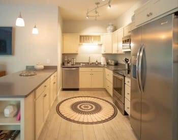 long view of kitchen with stanless steam appliances, counter with cream cabinets above and below, ceramic tiled floor with round rug by stove