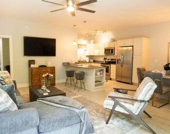 overview of 3 rooms together with gray couch, flat screen tv on wall, white chair, celing fan, kitchen in distance and dinining room with tiled floors