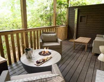 wooden deck with cream colored wicker chairs by round table with fire pit, wine in ice bucket, cheese tray, outside shower in background