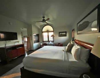 King bed with tan bedspread with striped throw pillows, two brown sitting chairs with a view of the mountains.