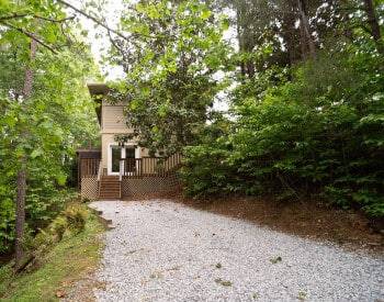 Gravel driveway in between trees on both sides leading to tan building with wodden deck with wide double doors