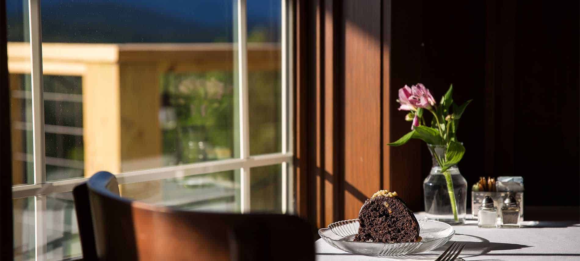 Chocolate cake on a glass plate, on a table with floral centerpiece, in a window with intentional focal blur.