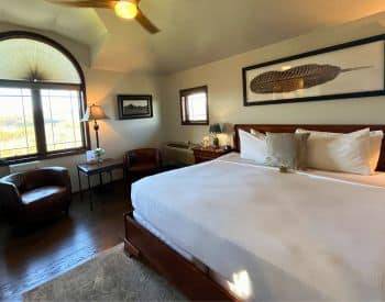 King bed with tan bedspread with striped throw pillows, two brown sitting chairs with a view of the mountains.