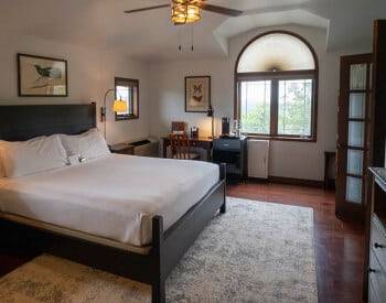 side view of king bed with white coverings on tan rug on hardwood floors, framed print over bed, desk with lamp on far wall in between two windows