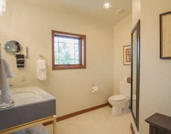 view toward window into corner of cream bathroom with sink on left and toilet in corner, long framed mirror on wall, stand on right with picture above