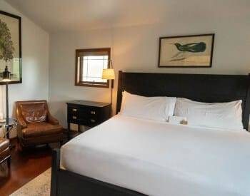 front view of king bed with white coverings with framed print over, sitting area with leather chairs and lamp with stand, end table by bed with floor lamp.