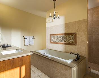 Tiled bathroom with warm colors and firm lines. A simple chandileer hangs over tub, with shower entry on side. 