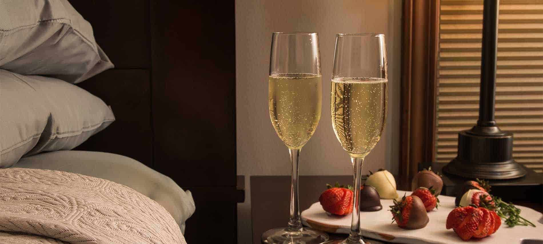 Two champagne glasses with gently bubbling liquid inside, stand next to chocolate covered strawberries on a bedside table.
