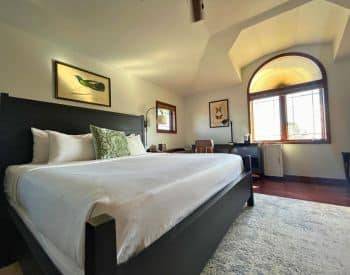 side view of king bed with white coverings on tan rug on hardwood floors, framed print over bed, desk with lamp on far wall in between two windows